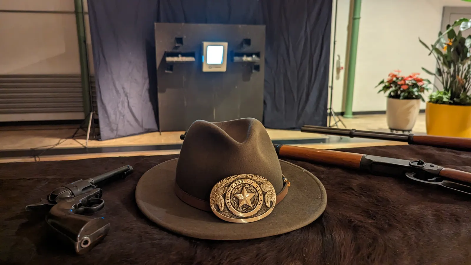 Cowboyhut, Revolver und Unterhebelgewehre auf dem Fell, dahinter die Zielwand